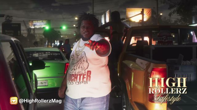 A heavyset Black man with dreadlocks and gold chains points at the camera, standing between a green car and…