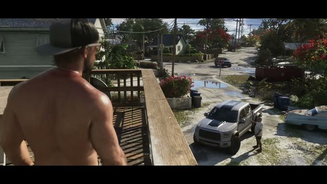 A shirtless man with a baseball cap stands on a wooden balcony overlooking a residential street with a…