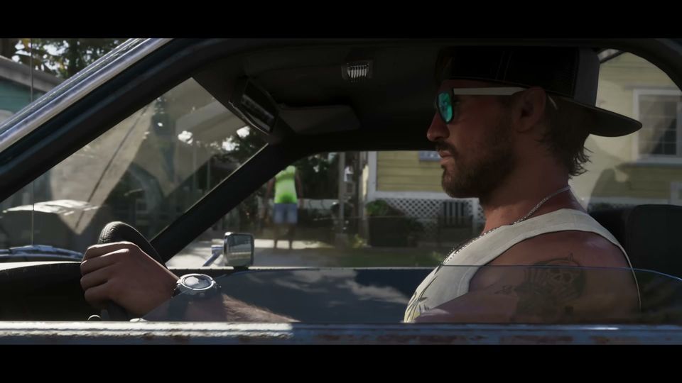 A man with a beard, sunglasses, and a baseball cap is seen from the side, driving a car with a tattoo visible on his left arm, while houses are visible outside the window.