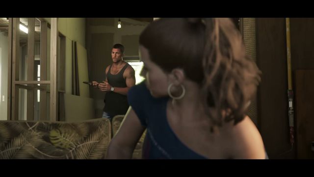 Lucia, out of focus in the foreground, looks away from Jason, who stands in the background holding a remote…
