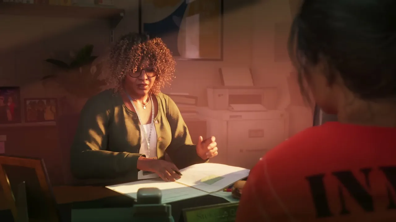 A woman with curly hair and glasses sits at a desk, speaking to another person whose back is…