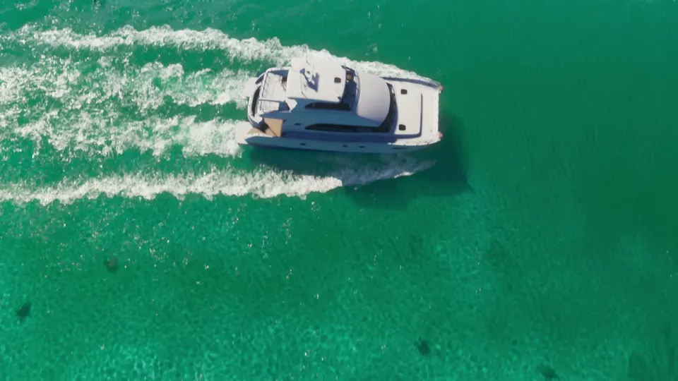 A white yacht is seen from an aerial perspective, cruising through clear turquoise water and leaving a white wake behind it.
