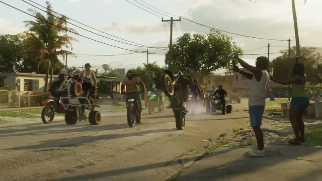 A group of people on dirt bikes and ATVs perform wheelies down a residential street while spectators watch…