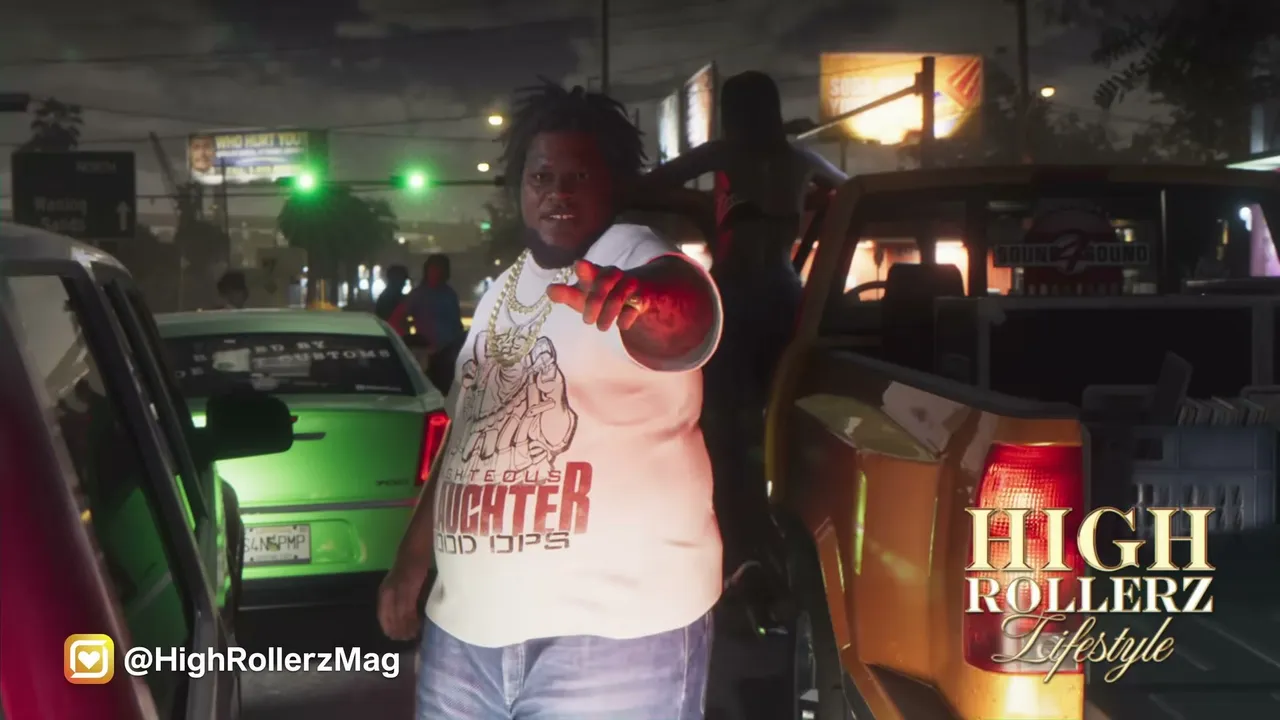 A heavyset Black man with dreadlocks and gold chains points at the camera, standing between a…