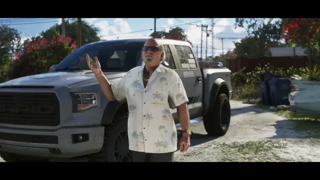 An older man wearing a tropical shirt and sunglasses stands next to a grey pickup truck, gesturing with his…
