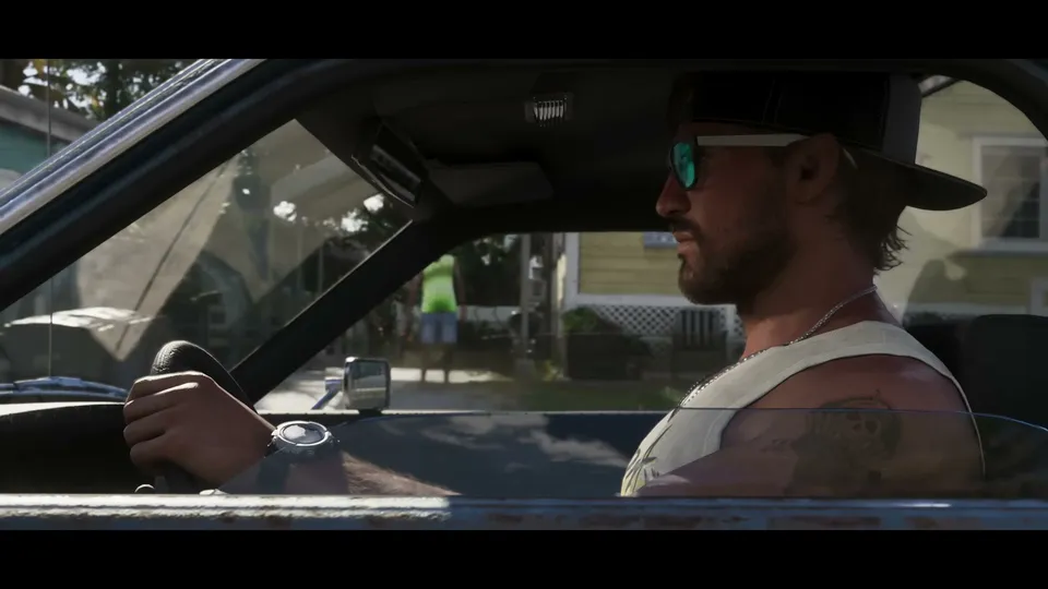 A man with a beard, sunglasses, and a baseball cap is seen from the side, driving a car with a tattoo visible on his left arm, while houses are visible outside the window.