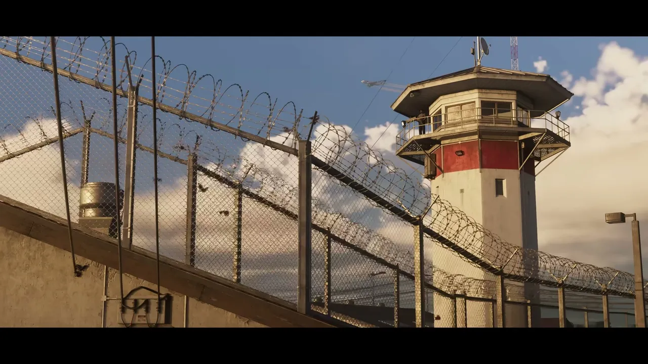 A prison watchtower with a guard visible stands behind chain-link fences topped with razor wire…