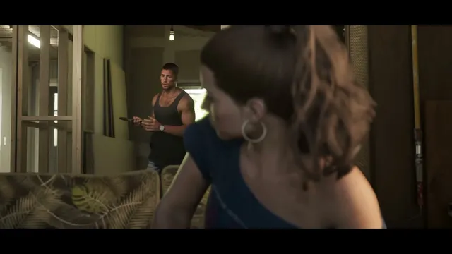 Lucia, out of focus in the foreground, looks away from Jason, who stands in the background holding a remote…
